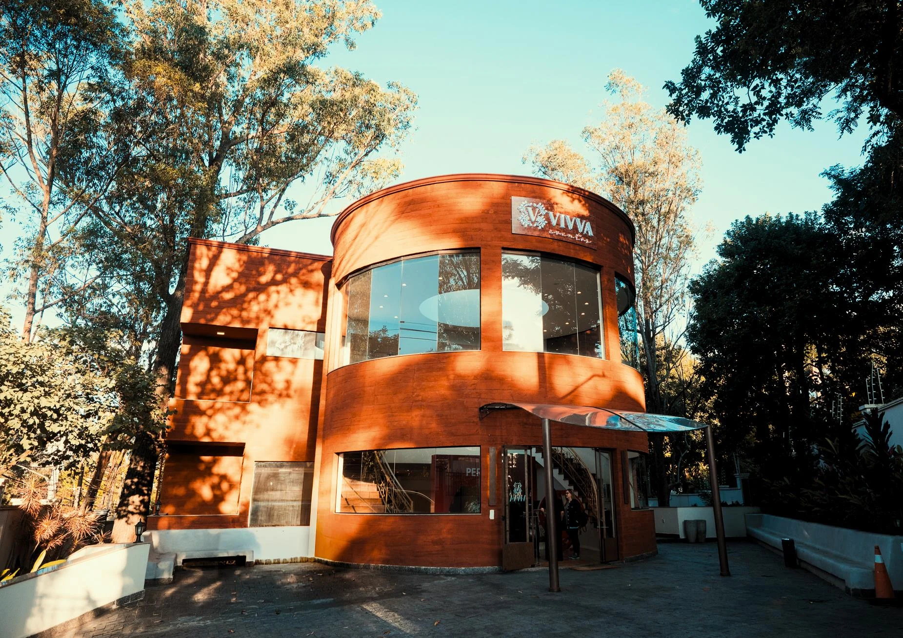 Fachada curva do Vivva Studio ao entardecer, entre as árvores
